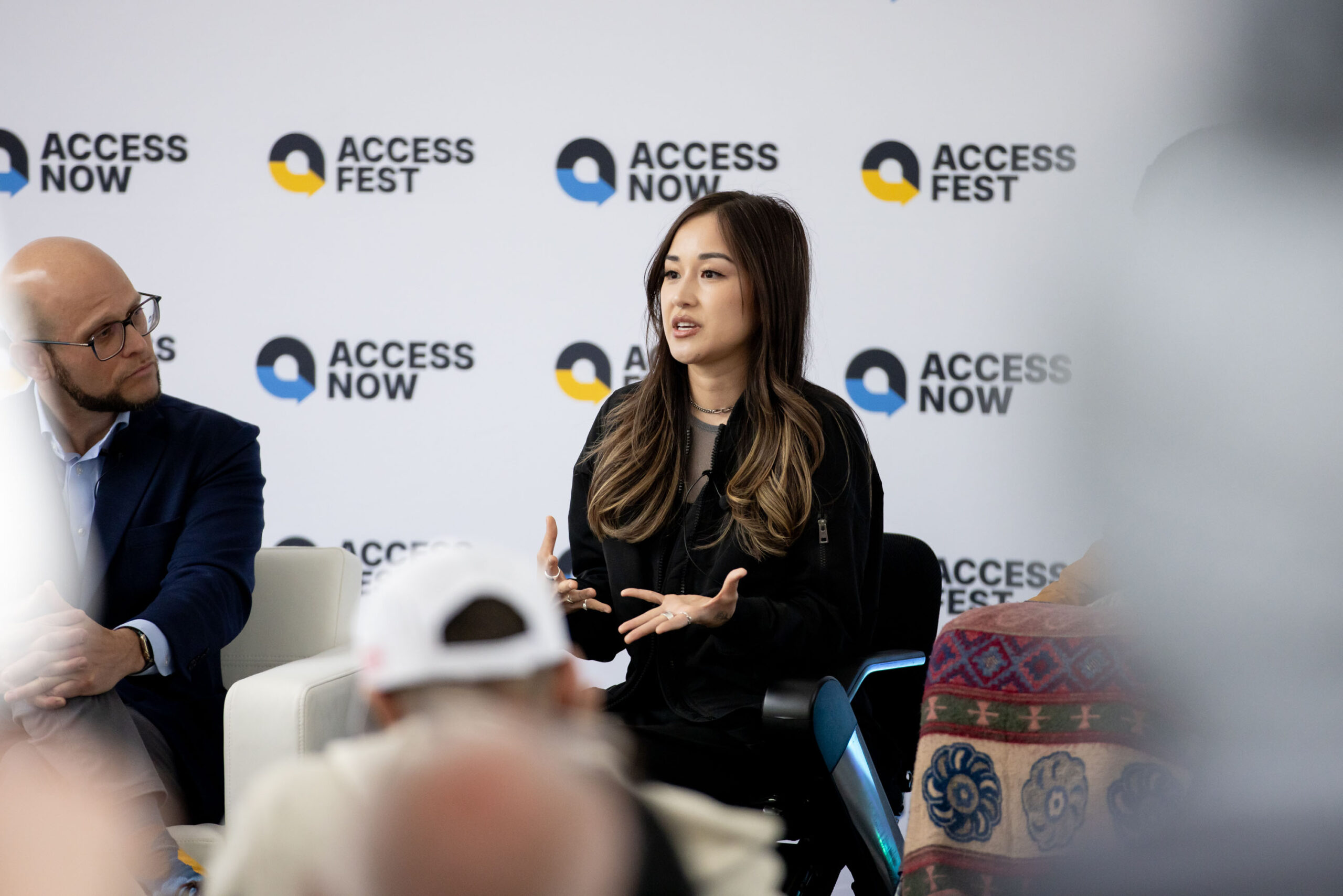 Panel discussion at AccessFest with three speakers on stage—one speaking, one in a wheelchair—while an audience listens, with AccessNow and AccessFest logos displayed on the backdrop.