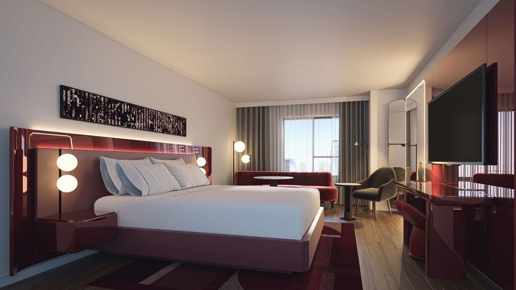 A modern hotel room with a king-sized bed featuring a red upholstered frame and built-in lighting, flanked by glowing round bedside lamps. The room includes a seating area with a red sofa, armchair, and small table near a window with floor-to-ceiling curtains, as well as a large flat-screen TV mounted across from the bed.