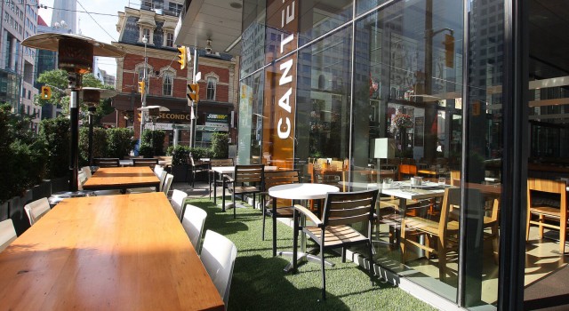 Outdoor patio seating at a modern restaurant with wooden tables and chairs arranged along a glass-walled building. A vertical sign reads “CANTEEN,” and nearby buildings and street details suggest a downtown urban setting.