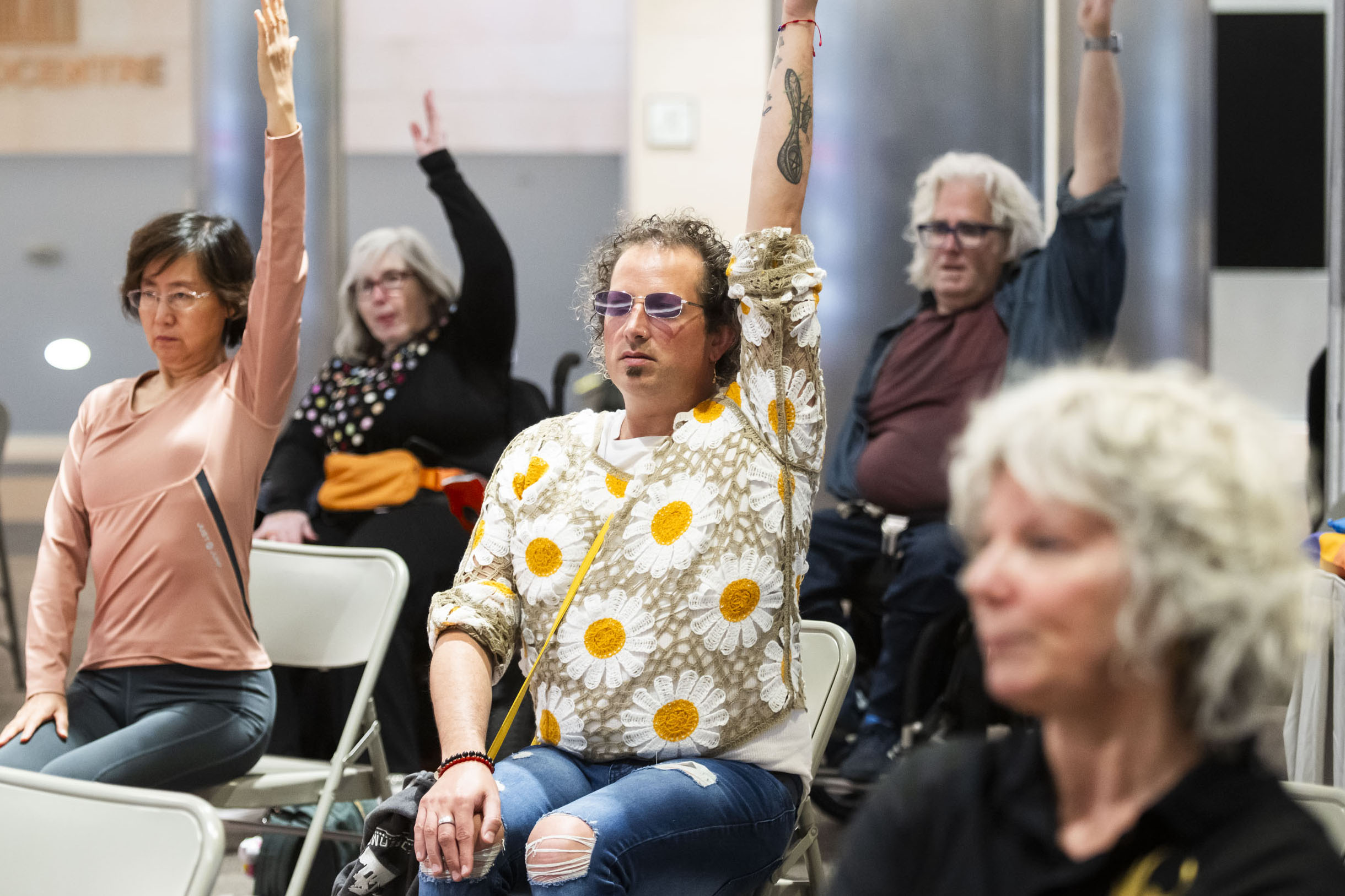 Participants seated in folding chairs take part in a guided movement or mindfulness session indoors, raising one arm overhead in unison. A diverse group of attendees, including older adults and people with disabilities, are spread throughout the room as they follow the exercise.