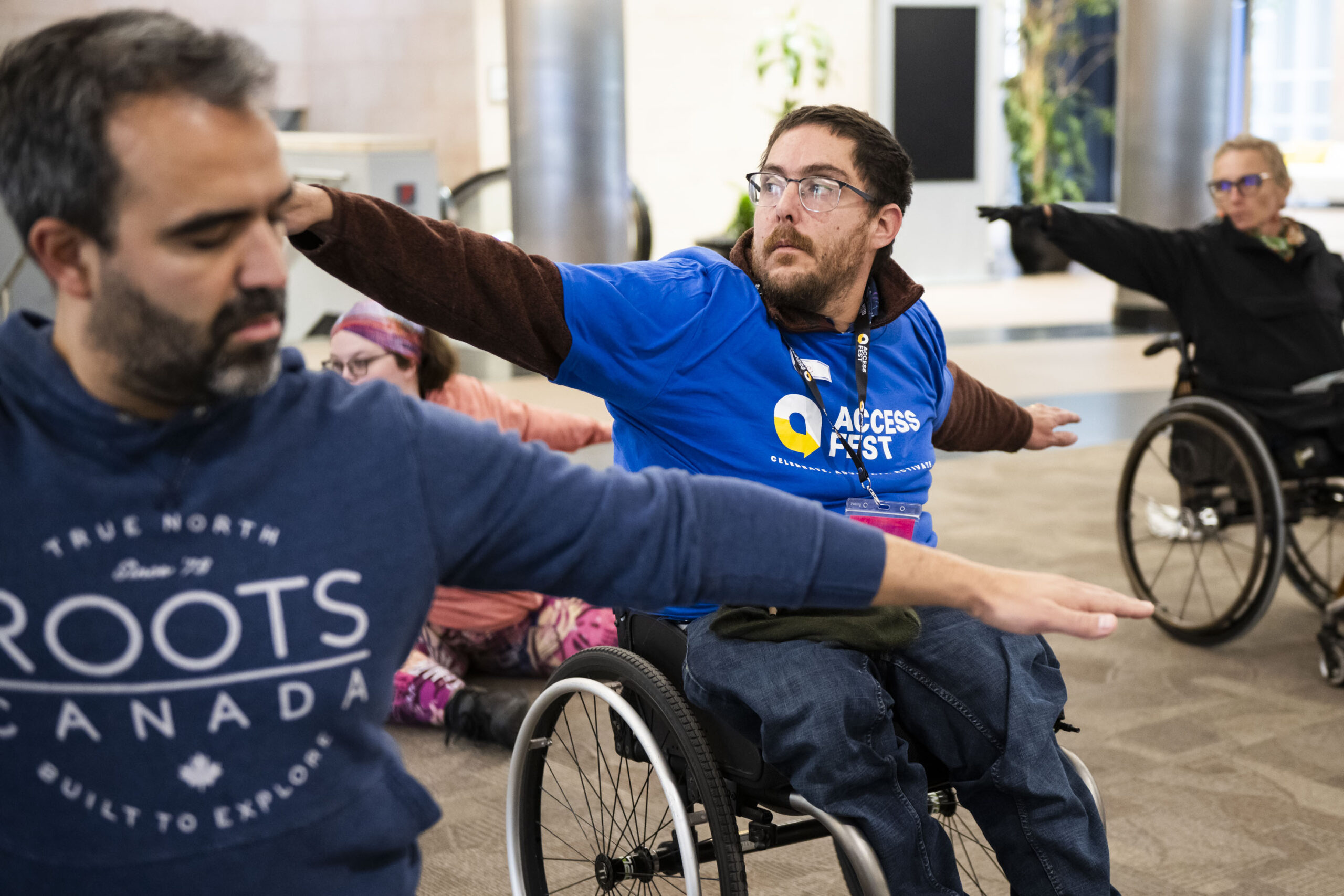 Participants take part in a seated movement or yoga session indoors, including wheelchair users and standing attendees stretching their arms outward while following the activity leader at AccessFest.
