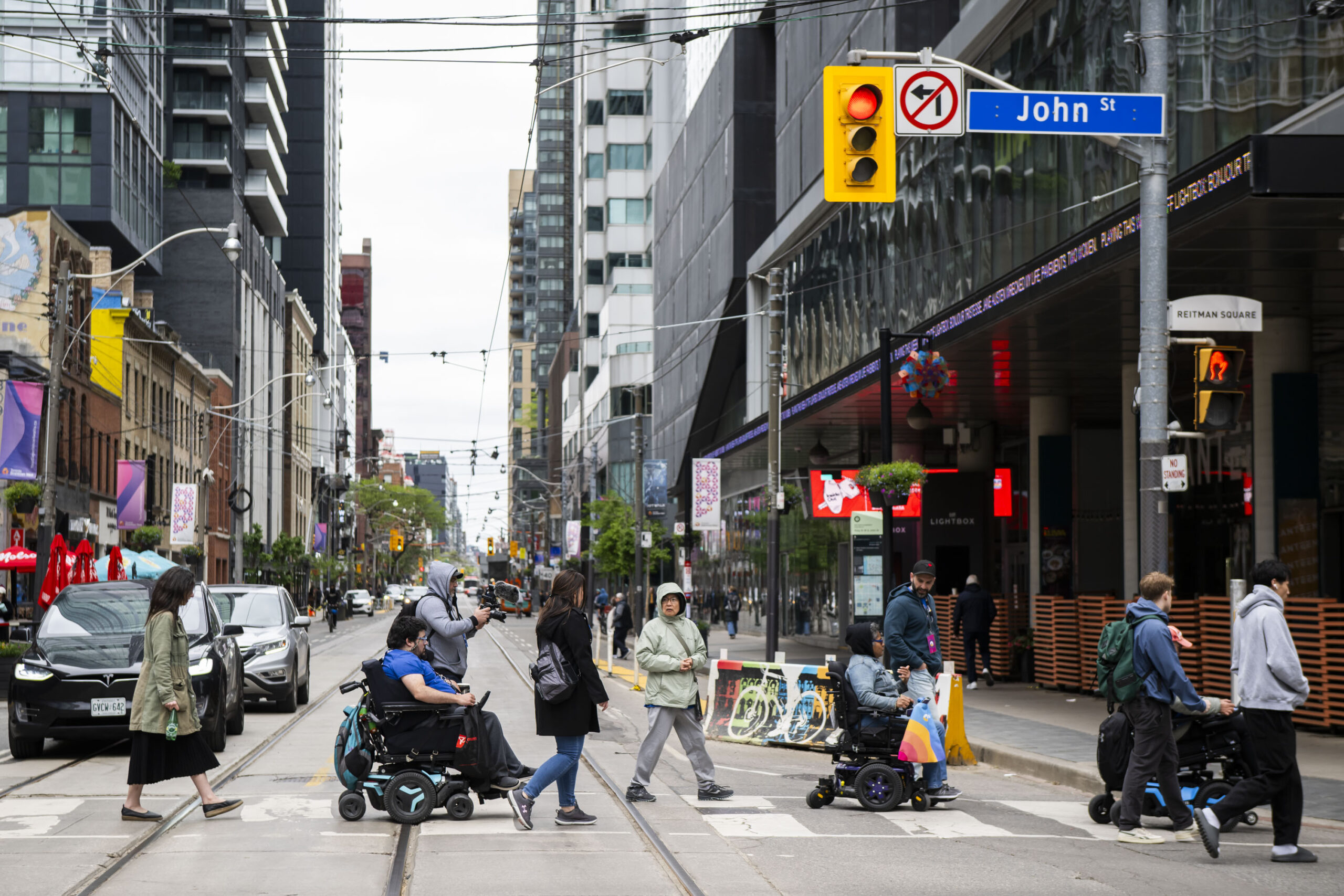 People cross King Street West at John Street in downtown Toronto, including several power wheelchair users, while others walk alongside them through a busy urban intersection lined with condos, restaurants, and streetcar tracks.