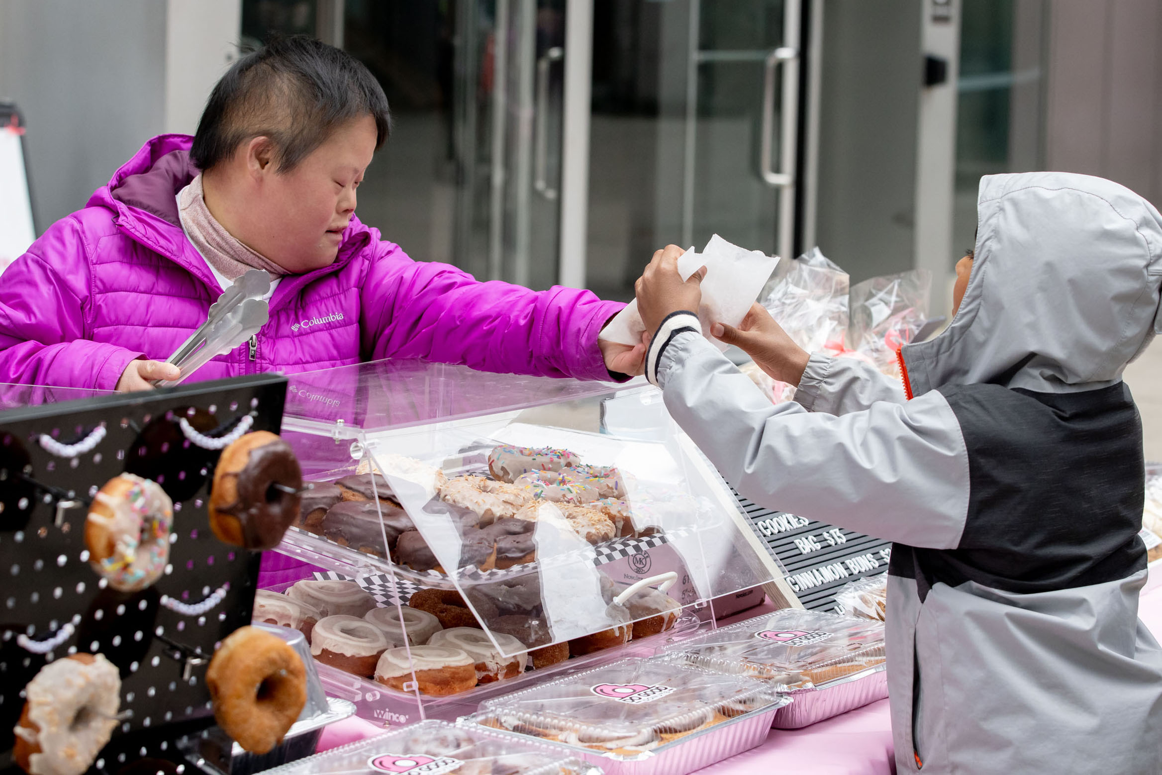 A person in a bright purple jacket reaches across a donut vendor table to receive a paper bag from a child in a gray jacket. Trays of assorted donuts and baked goods are displayed in clear cases between them, creating a warm moment of exchange and community at an outdoor festival market.
