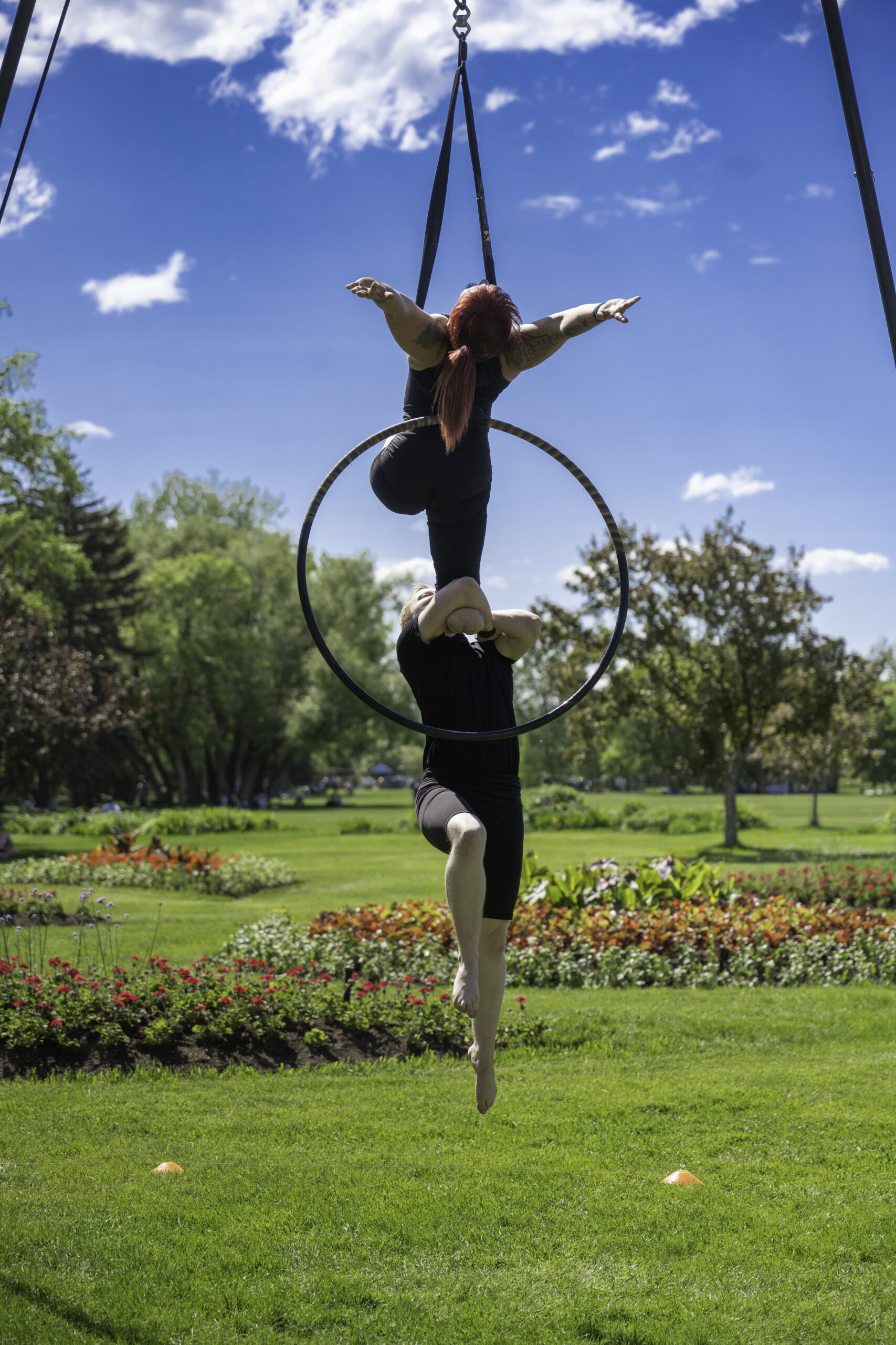 Two aerial performers balance together on a suspended hoop in a park on a sunny day. One performer, Maxime, balances within the hoop while supporting the other above them, Erin, who extends their arms outward against a bright blue sky. Colourful flower gardens and green lawns surround the performance space.