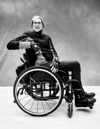 Black-and-white portrait of a woman seated sideways in a manual wheelchair against a textured studio backdrop. She wears glasses, a knit beanie, fingerless gloves, a quilted vest layered over a long-sleeve top, jeans, and ankle boots. Resting one arm casually on the wheelchair’s backrest, she faces the camera with a warm smile and relaxed posture. The wheelchair is shown in profile, with its large rear wheel prominently visible. The portrait has a confident, outdoorsy feel, highlighting both her personality and independence in a poised and approachable way.