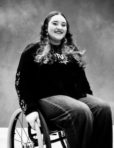 Black-and-white portrait of a smiling young woman seated in a manual wheelchair against a textured studio backdrop. She has long wavy hair with lighter ends and wears a dark cardigan over a T-shirt and wide-leg jeans. One hand rests lightly on the wheelchair’s push rim while she faces the camera with a warm, confident expression. The wheelchair is partially visible, framing her seated posture without dominating the image. The portrait feels relaxed and welcoming, highlighting her personality, self-assurance, and presence.