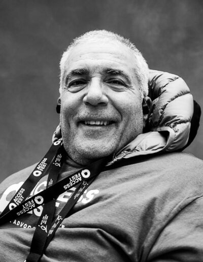 Black-and-white portrait of an older adult with short gray hair smiling warmly toward the camera. They are seated and wearing a sweatshirt with an AccessFest lanyard draped across their chest. A padded neck support wraps around the back of their head, and a soft, neutral studio backdrop keeps the focus on their relaxed expression and welcoming presence.