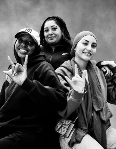 Black-and-white portrait of three smiling young women posed closely together against a textured studio backdrop. Two sit in the foreground while one stands behind them with her arms resting across their shoulders. The woman on the left wears a baseball cap and hoodie, while the woman on the right wears a headscarf and layered casual clothing. Both hold up hand gestures and smile toward the camera. The woman standing behind them wears hoop earrings and a zip-up jacket, looking directly at the camera with a relaxed expression. The portrait conveys friendship, confidence, and community, with the group appearing comfortable, playful, and connected.