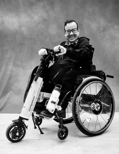 Black-and-white portrait of a smiling man seated in a manual wheelchair fitted with a front-mounted power-assist attachment. He faces slightly toward the camera with both hands resting on the handlebars of the device, creating a dynamic, ready-to-go pose. He wears glasses, a dark jacket, dark pants, and sneakers. The wheelchair’s large rear wheels and the rugged front attachment are clearly visible, highlighting adaptive mobility technology.
