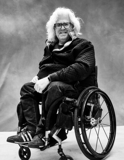 Black-and-white portrait of a person seated in a manual wheelchair against a textured studio backdrop. They have shoulder-length light-coloured hair, wear glasses, and are dressed in a quilted jacket, dark jeans, and sneakers. Their hands rest comfortably in their lap as they face the camera with a relaxed, friendly expression. The wheelchair’s sleek frame and large rear wheels are visible in profile. The portrait has a calm, confident feel, emphasizing personality, presence, and self-assurance.