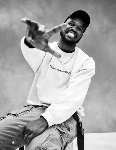 Black-and-white portrait of a smiling man seated casually on a stool against a textured studio backdrop. He wears a baseball cap, a light-coloured sweatshirt, cargo pants, and a chain necklace. Leaning slightly forward, he reaches one hand playfully toward the camera, creating a sense of movement and connection. His wide smile and relaxed posture convey confidence, warmth, and joy. The close perspective emphasizes his expressive gesture and energetic personality, giving the portrait a lively and engaging feel.