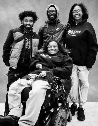Black-and-white group portrait of four smiling young adults posed against a textured studio backdrop. One person sits at the front in a power wheelchair, wearing glasses and a dark hoodie, while three others stand behind them. The group is dressed casually in hoodies, jackets, and athletic wear. Two people wear matching AccessFest hoodies, and all four look directly toward the camera with warm, confident expressions. The portrait conveys friendship, community, and a sense of belonging, highlighting a diverse group gathered together in a relaxed and welcoming setting.