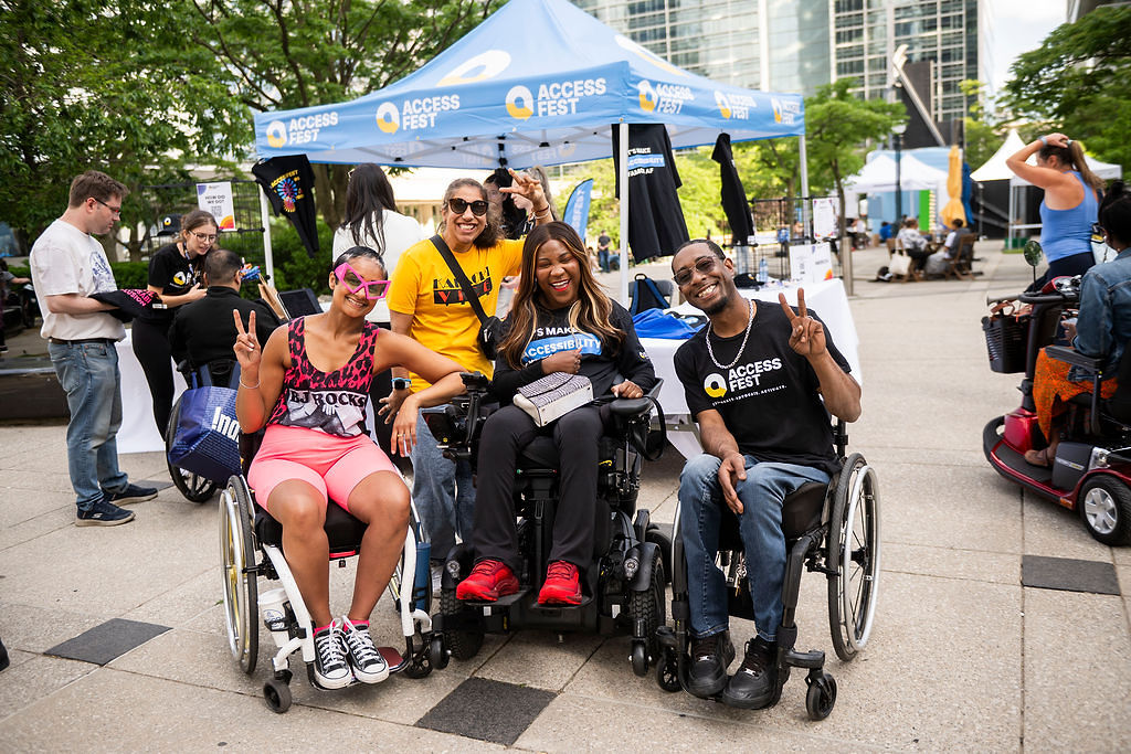 Four individuals smiling and doing the peace signs with their hands, at AccessFest 2026. Three of the individuals are using wheelchairs, and one person is standing behind them.
