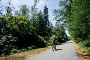 a person biking down a wide and paved trail, surrounded by forestry