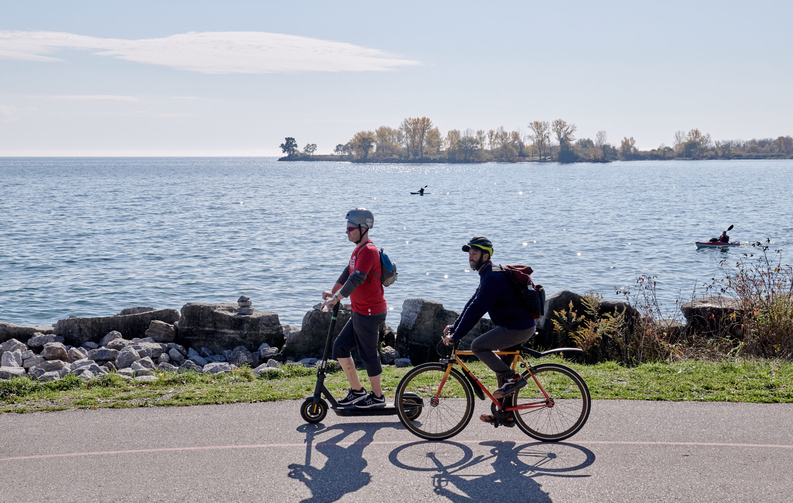 two accessnow mappers on the martin goodman trail, one person is on a scooter and another person is on a bike. Behind them is the water and the skyline.