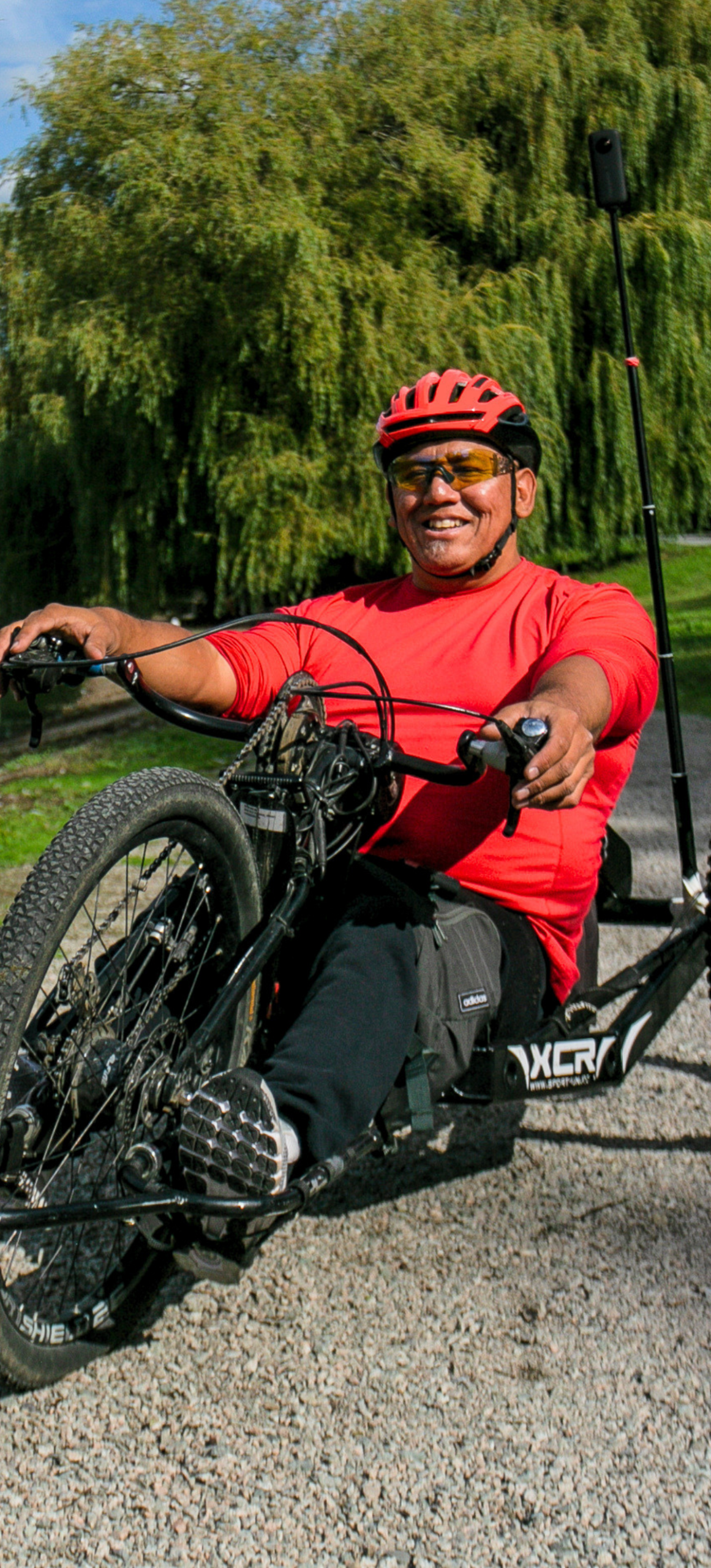 A smiling man wearing a red shirt, red bicycle helmet, and sunglasses rides a recumbent adaptive handcycle along a gravel path, surrounded by lush green trees.