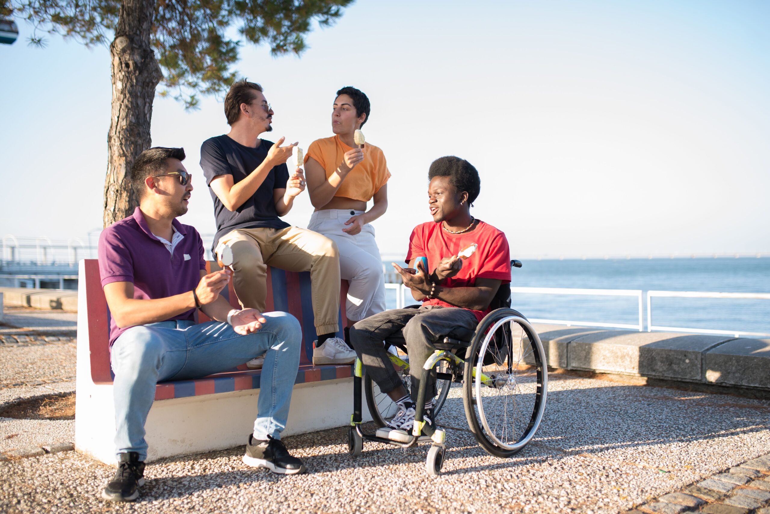A group of friends sitting outside. They are engaging in conversation and enjoying ice cream. One person is seated on a wheelchair, others are seated on a bench.