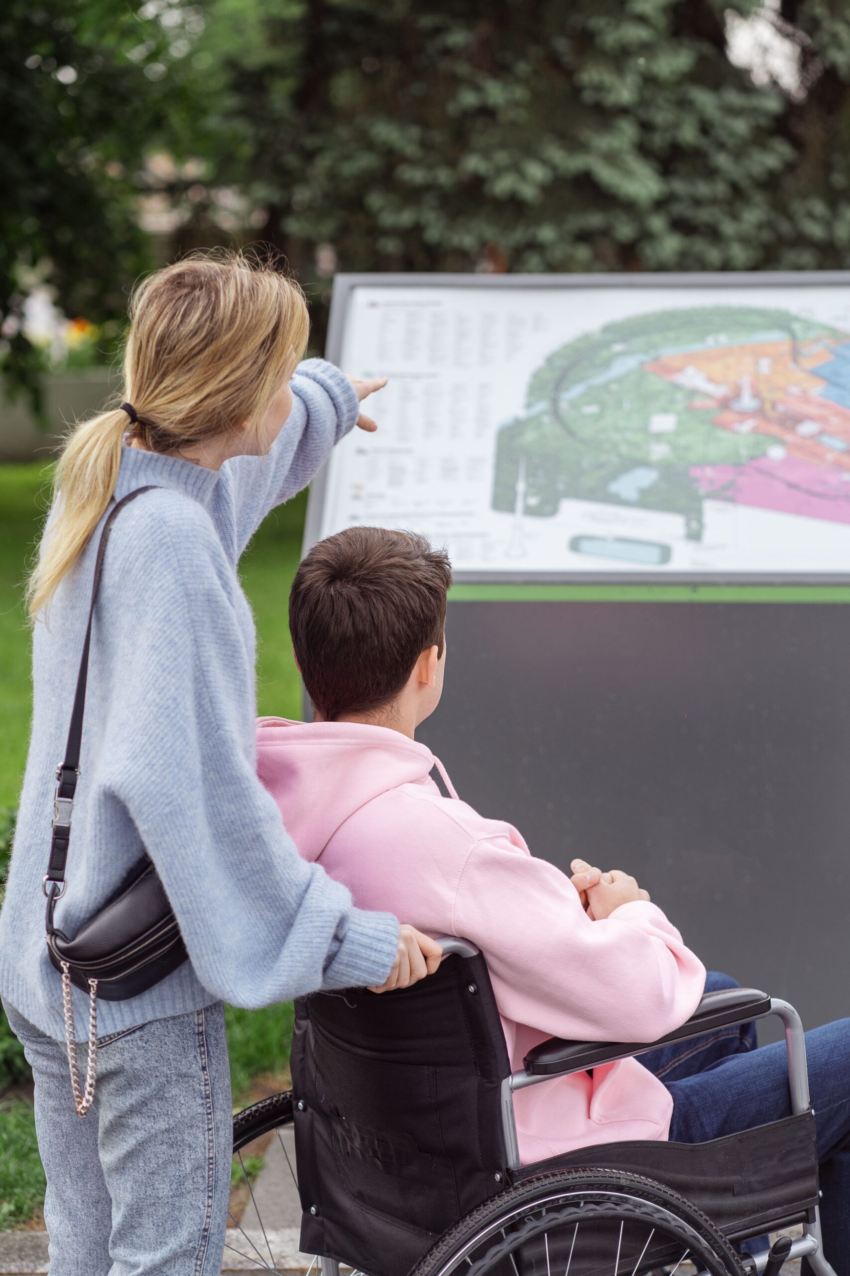 a female pushing a wheelchair with a male seated in the wheelchair, outdoors. they have stopped and are looking at a map she is pointing at. 