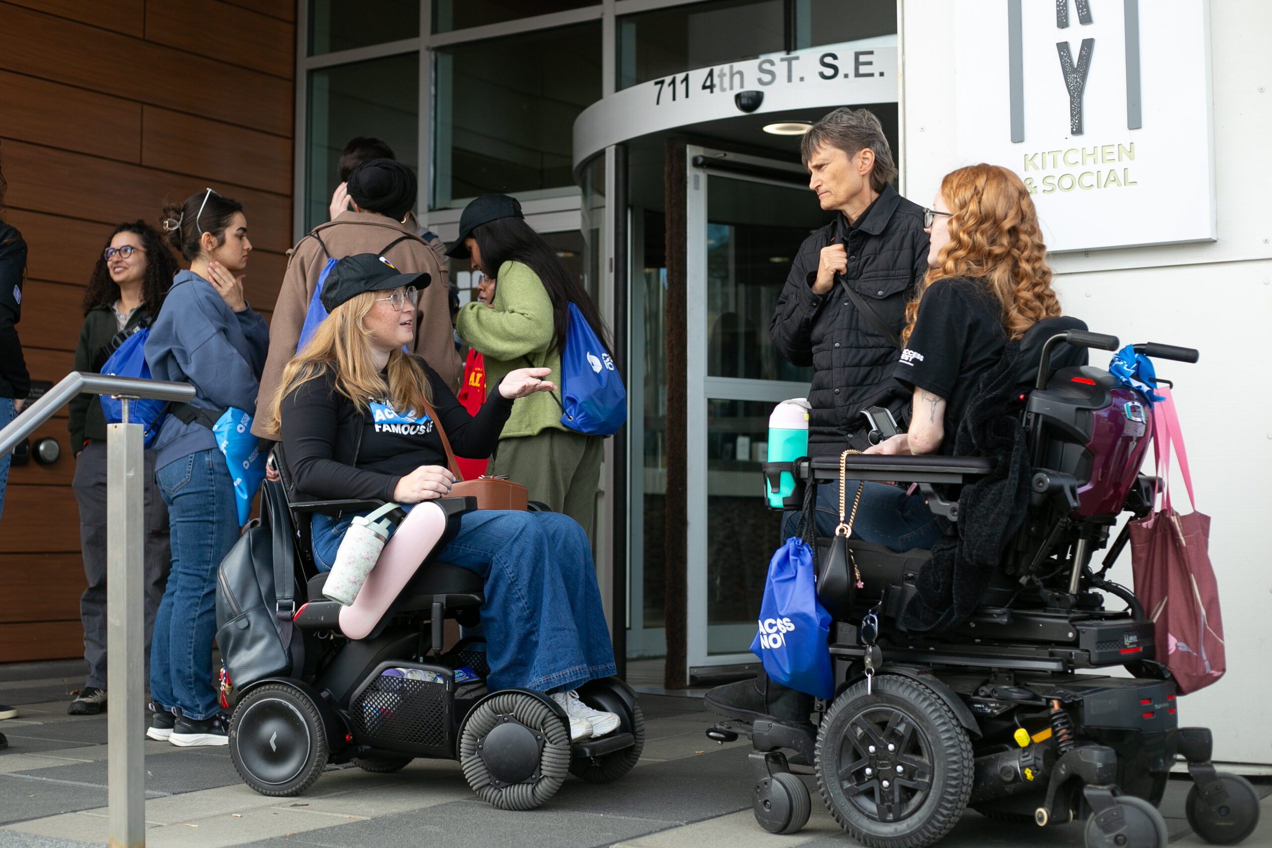 Two women in power wheelchairs chat with a third person standing between them outside the entrance of a building at 711 4th St. S.E. One wears a black cap and glasses and gestures as she talks. Behind them, a group of people with blue AccessNow drawstring bags gather near the doors.