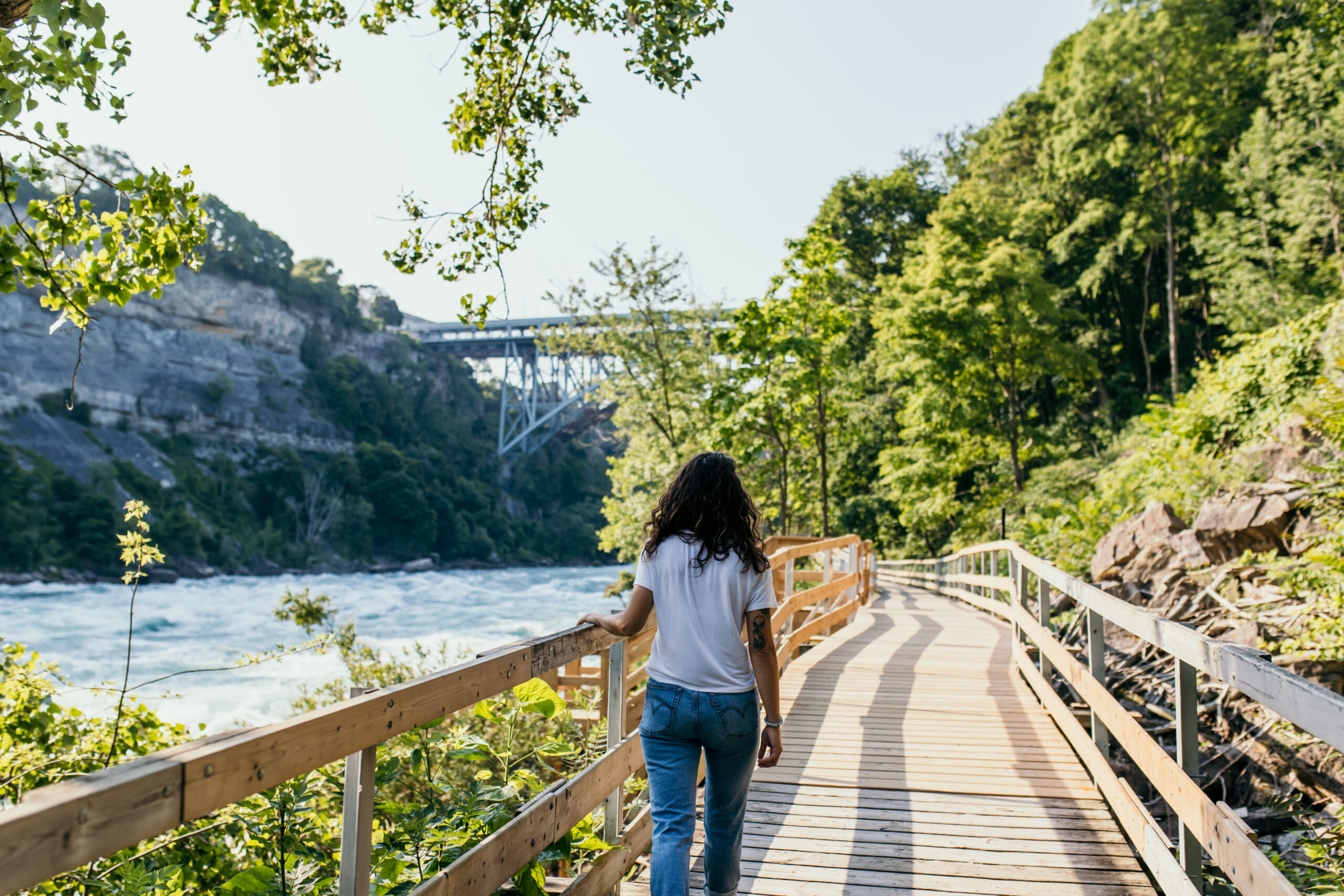 Woman walking along a wooden riverside boardwalk, surrounded by green trees, with a bridge in the distance.